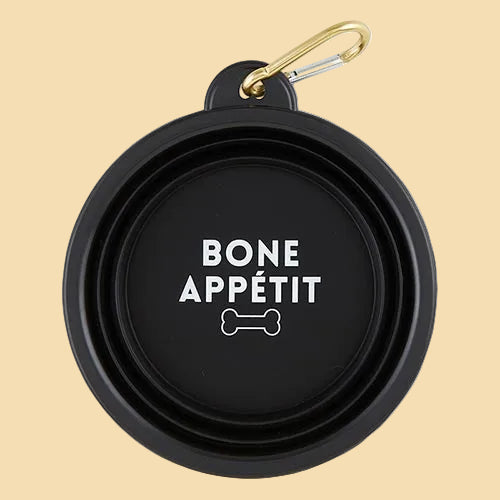 A black collapsible dog bowl with a gold carabiner clip, featuring the text 'BONE APPÉTIT' and a bone icon printed in white on the inside of the bowl,.