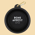 A black collapsible dog bowl with a gold carabiner clip, featuring the text 'BONE APPÉTIT' and a bone icon printed in white on the inside of the bowl,.