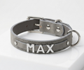 A gray faux-leather dog collar with silver hardware, featuring the name 'MAX' in raised silver letters on the front, displayed on a white block against a white background.