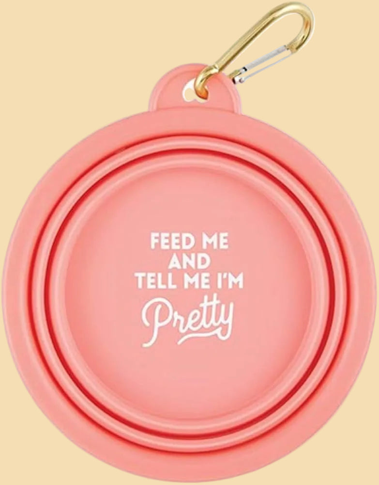 A coral pink collapsible pet water bowl with white printed text reading 'FEED ME AND TELL ME I'M Pretty' and a gold carabiner attached at the top, shown on a white background.